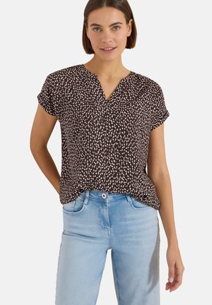 Femme portant un chemisier noir à manches courtes avec des taches blanches et un jean bleu clair, debout avec une main derrière le dos.