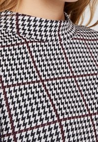 Donna che indossa un top a collo alto con un motivo a scacchi bianco e nero sovrapposto da sottili linee di griglia rosso scuro.