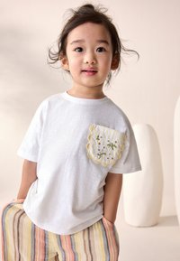 Fehér pamut póló sárga, hullámos szegélyű zsebbel, virágos hímzéssel díszítve. Többszínű csíkos nadrággal párosítva.