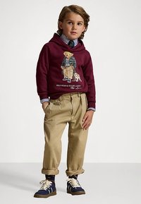 Niño pequeño con un hoodie marrón con gráfico del oso Polo, camisa a rayas, corbata, pantalones beiges y zapatillas navy, de pie con las manos en los bolsillos.