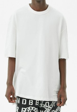 Hvid oversized t-shirt med korte ærmer, lavet af blødt bomuld. Har frynset kanter og et lille mærke på siden.