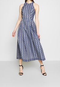 Blå och vit rutmönstrad midiklänning med halterneck, åtsittande liv och utsvängd kjol. Slät textur, minimalistisk design, kombinerad med svarta klackar.