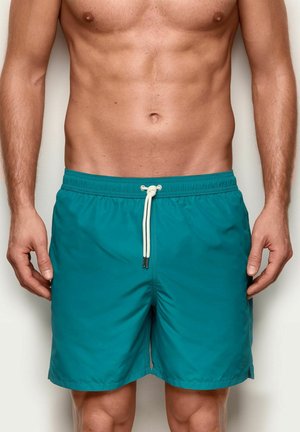 Męski tors w turkusowych kąpielówkach z białym troczkiem, stojący z ramionami luźno opuszczonymi wzdłuż boków na jednolitym tle.