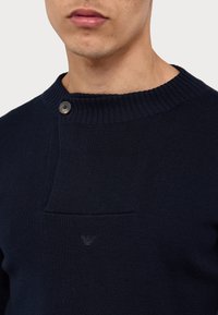 Gros plan d’un homme portant un pull en maille bleu marine avec un col côtelé, un patch carré, un bouton unique et un logo brodé discret.