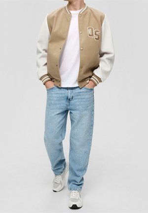 Personne portant une veste universitaire beige et blanche avec un patch "05", un t-shirt blanc, un jean bleu clair et des baskets blanches, les mains dans les poches.