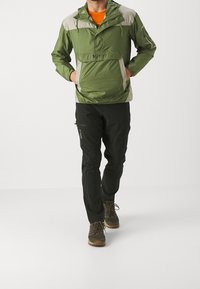 Chaqueta tipo pulóver verde con detalles en gris claro, bolsillo frontal y capucha ajustable. Combinada con pantalones negros y botas de senderismo marrones.