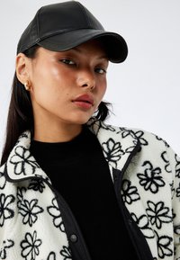 Giacca di pile bianca con motivi floreali neri, indossata sopra un maglione nero. Un berretto di pelle nera e orecchini a cerchio dorati completano il look.