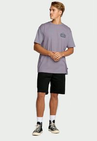 Jonge man staand in een lila T-shirt met een kleine opdruk, zwarte shorts, witte sokken en zwarte hoge sneakers, kijkend naar rechts.