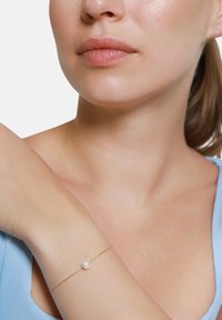 Gulltrådsarmbånd med en enkelt hvit perle, båret på en bar arm. Armbåndet er delikat med et minimalistisk design.