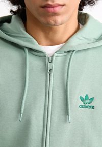 Casaco com fecho éclair Adidas de verde claro, com tecido texturizado, cordões ajustáveis e logótipo bordado no lado esquerdo do peito.