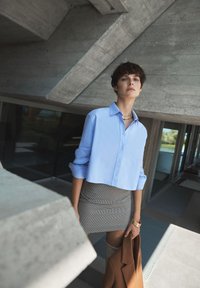 Camicia azzurra chiara a maniche corte con maniche arrotolate, abbinata a una gonna grigia con motivo. Si tiene un cappotto marrone e un braccialetto dorato.