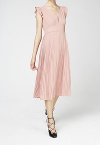 Frau in einem rosa plissierten Midikleid mit Rüschensäumen und schwarzen Schnür-Heels, die vor einem schlichten weißen Hintergrund steht.