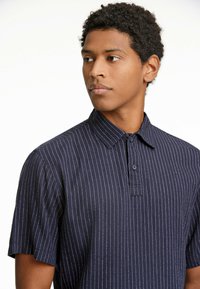 Marinblå kortärmad pikétröja i strukturerad tyg med vertikala vita ränder, med en knappad krage och en framficka.