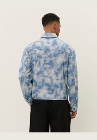 Light blue denim jacket with a cloud-like pattern, buttoned cuffs, and a classic collar. Textured surface with a slightly oversized fit.