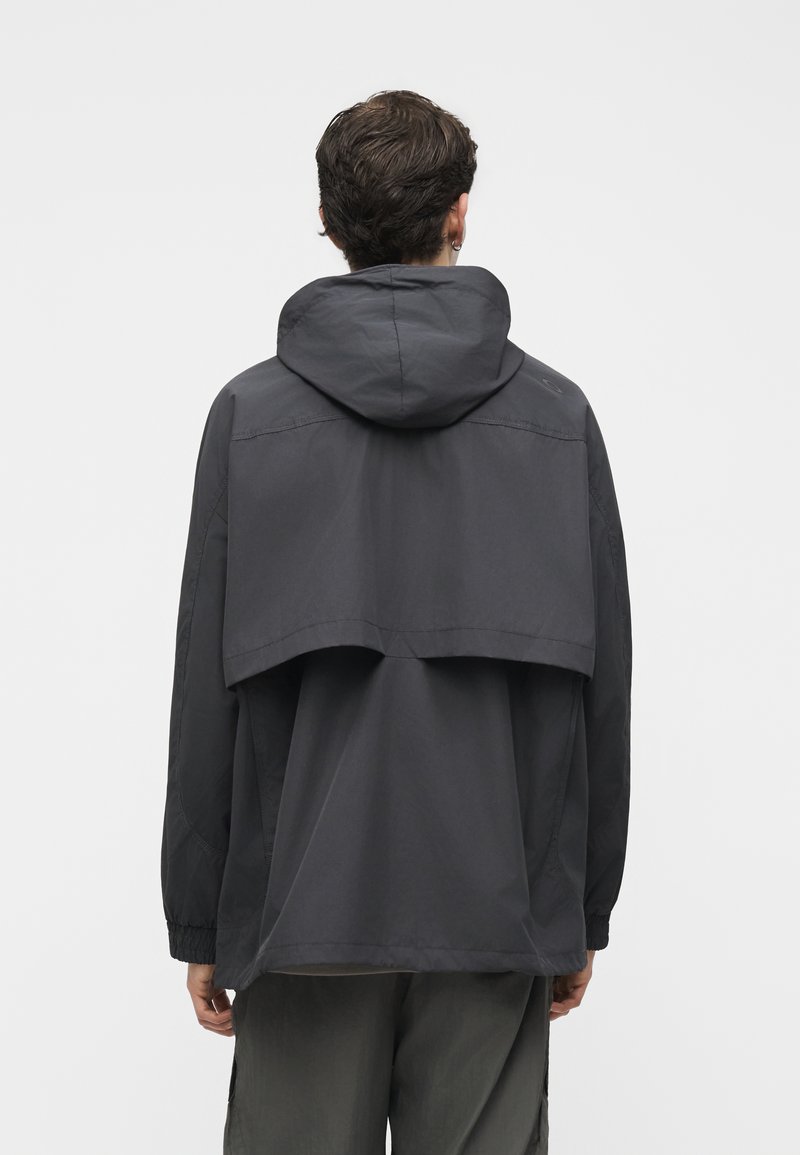 Hombre que lleva una chaqueta con capucha de color gris oscuro con un detalle en la parte trasera parecido a una capa, de pie mirando hacia atrás contra un fondo liso.