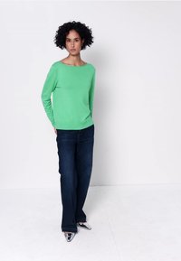 Femme aux cheveux bouclés portant une chemise verte à manches longues, un jean large bleu foncé et des chaussures argentées pointues, debout contre un mur blanc.