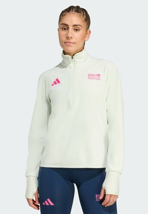 Donna che indossa una felpa a zip corta verde chiaro e leggings blu navy con i loghi rosa del BMW Berlin Marathon e di Adidas, in piedi contro uno sfondo semplice.