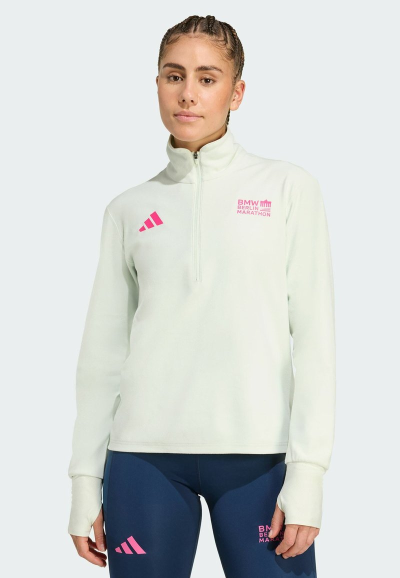 Žena v světle zelené mikině s krátkým zipem a tmavě modrých legínách s růžovými logy BMW Berlin Marathon a Adidas, stojící na jednobarevném pozadí.