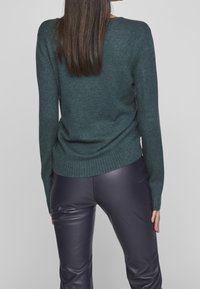 Pull vert foncé en maille avec ourlet côtelé, associé à un pantalon en simili cuir noir, vu de dos.