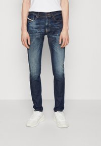 Los jeans de mezclilla azul oscuro tienen un corte ajustado, un ligero desgaste y un sutil efecto de distress. Combinados con zapatillas blancas, el conjunto ofrece un aspecto casual.