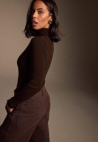 Suéter de cuello alto acanalado color marrón combinado con pantalones de talle alto en marrón oscuro. La modelo lleva pendientes grandes de tono dorado. Fondo discreto.