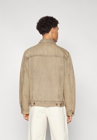 Beige denim jakk, millel on nööpidega esiosa, õmmeldud detailid ja nööpide aksessuaarid mansettides, kujutatud tagant.