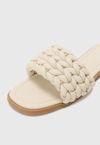 Sandale en cuir couleur crème avec une large sangle tissée, un dessous de pied en cuir lisse et une semelle en caoutchouc marron clair. Design minimaliste et détails texturés.