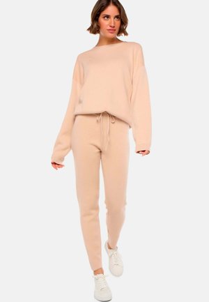 Une femme marche vers la caméra, portant un pull beige et un pantalon assorti, complétés par des baskets blanches et une coiffure décontractée.