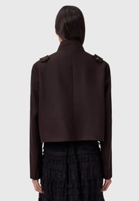Chaqueta corta marrón con botones en los hombros y un cuello estructurado, hecha de una tela texturada, que combina con una falda negra con capas.