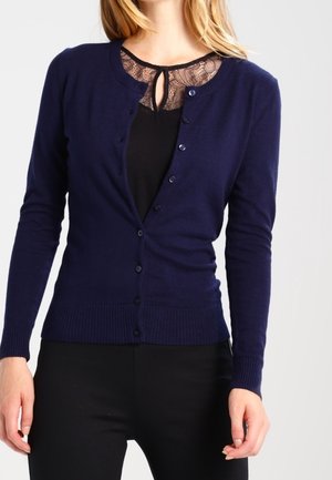 Nainen pukeutuneena istuvaan tummansiniseen napitettuun neuletakkiin mustan topin päällä, jossa on pitsinen detalji pääntiessä, yhdistettynä mustiin housuihin.