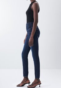 Jeans de mezclilla azul de cintura alta con un corte ajustado, que presentan bolsillos traseros y un ligero desgaste. Combinados con una blusa sin mangas de canalé negra y sandalias negras.