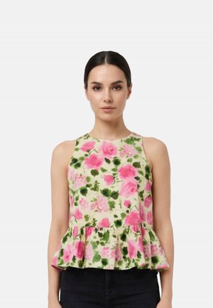 Donna con top beige senza maniche con stampa floreale rosa e verde e orlo a peplo, abbinato a pantaloni neri, in piedi davanti a uno sfondo bianco.