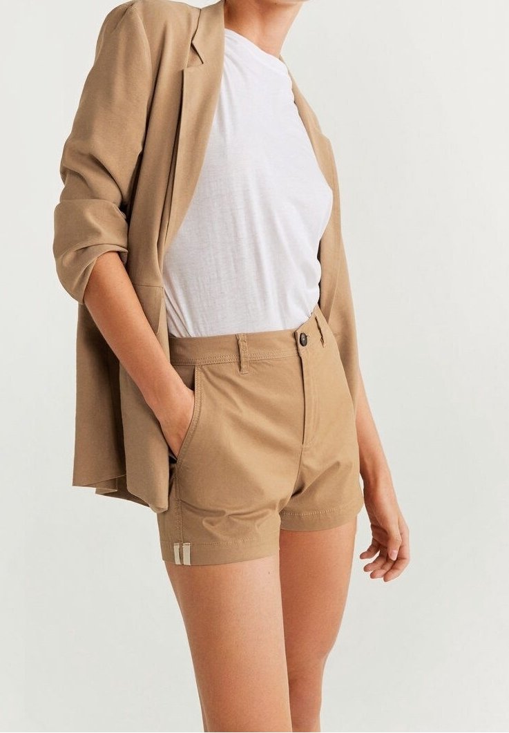 Femme portant un blazer beige et un short taille haute assorti avec un t-shirt blanc, debout avec une main dans la poche sur un fond uni.