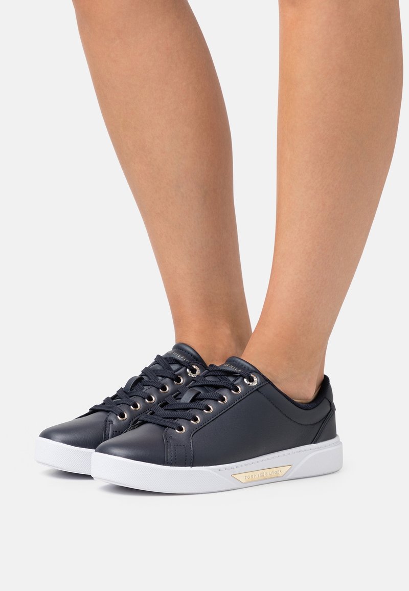 Tmavě modré kožené tenisky s bílými gumovými podrážkami, kovovými očky a texturovanými tkaničkami. Na boku mají zlatý detail.