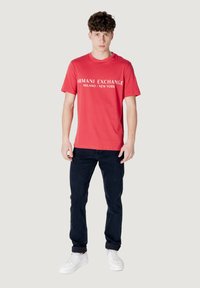 T-shirt in cotone rosso con stampa "ARMANI EXCHANGE MILANO / NEW YORK" in bianco, abbinata a jeans blu scuro e sneakers bianche.