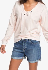 T-shirt long à manches en rose clair, côtelé, avec un décolleté en V et des détails à laçage. Associé à un short en jean, avec des manches retournées et une coupe classique.