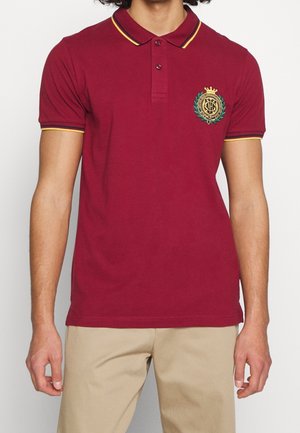 Homme portant un polo bordeaux à manches courtes avec des bordures jaunes et noires et un écusson brodé sur la poitrine, associé à un pantalon beige.