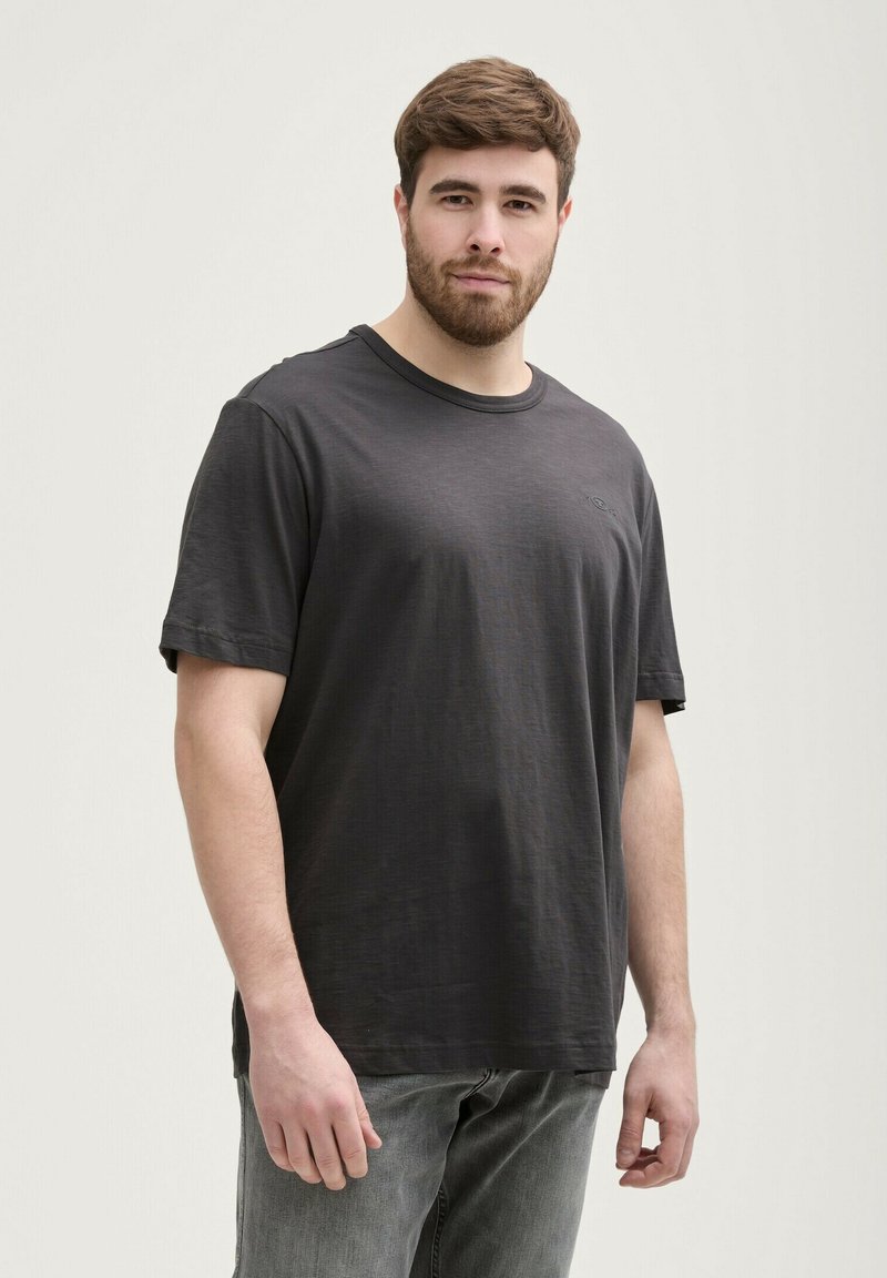 T-shirt noir à manches courtes avec un col rond et un logo discret. Texture douce, coupe décontractée, associée à un jean gris clair.