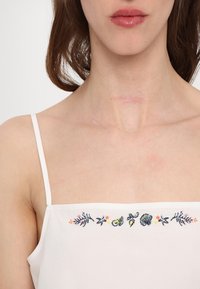 Hvit singlet med tynne stropper, med flerfarget blomstermønster og skjellbroderi langs halslinjen og en glatt bomullsstruktur.