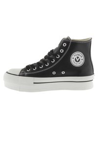 Zapatillas alta negras de material suave, con puntera y suela de goma blanca, cordones negros y un logo circular en el lateral.