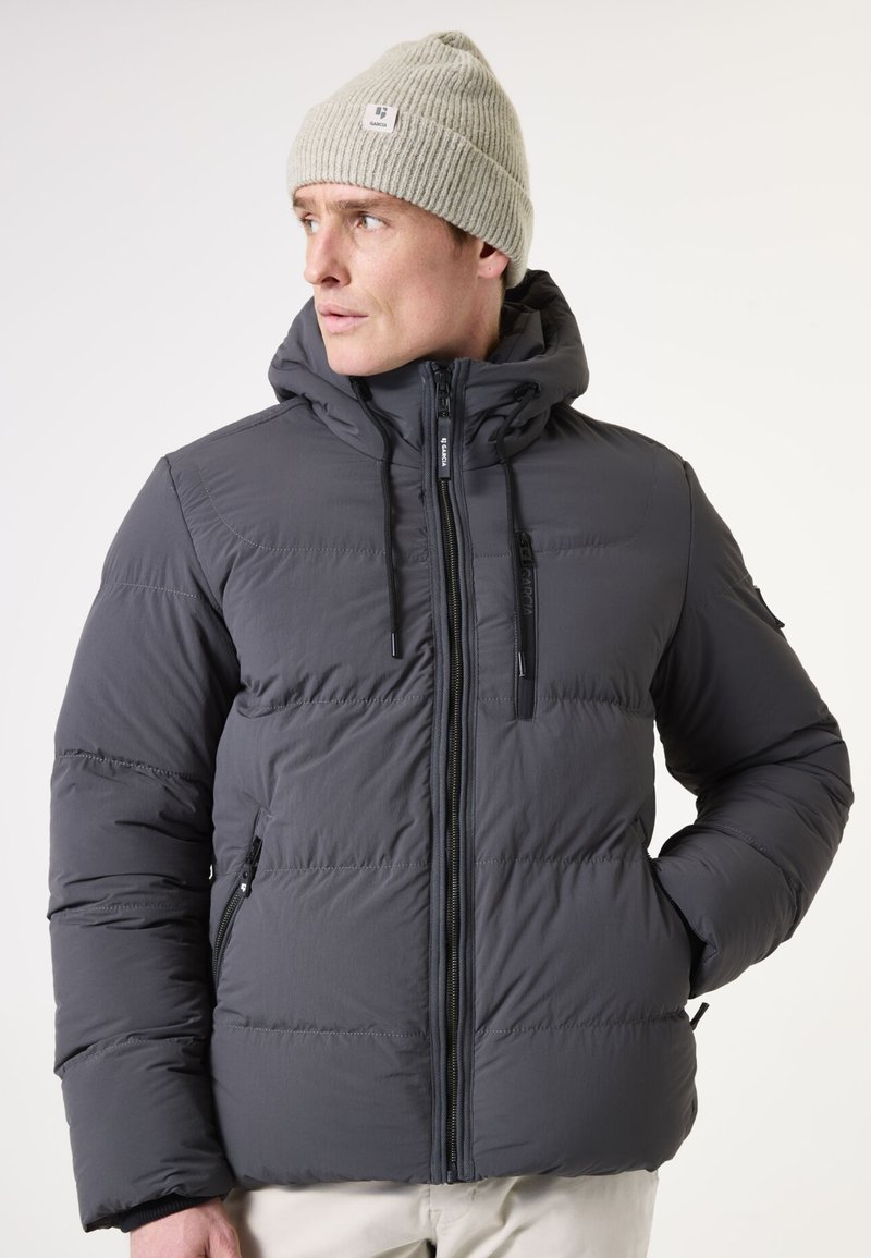 Veste rembourrée grise avec capuche, présentant un design quilté horizontal, fermeture éclair à l'avant et cordons réglables. Portée avec un bonnet léger.