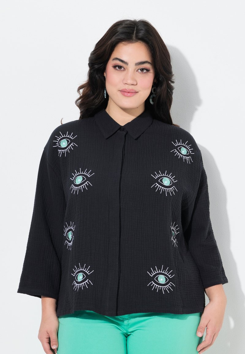 Zwarte button-up shirt met geborduurde oogontwerpen in teal en wit; gestructureerde stof en driekwartmouwen. Gecombineerd met mintgroene broeken.