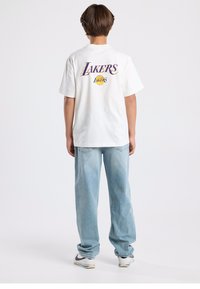 White t-shirt with "LAKERS" graphic in purple and yellow on the back, paired with loose-fitting light blue jeans and white sneakers.