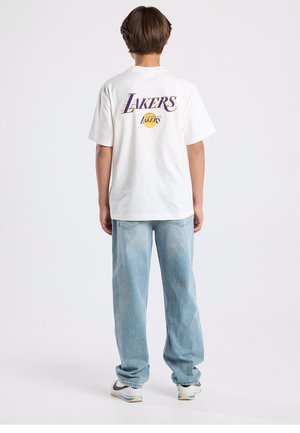Camiseta blanca con gráfico "LAKERS" en púrpura y amarillo en la parte trasera, combinada con vaqueros holgados de color azul claro y zapatillas blancas.