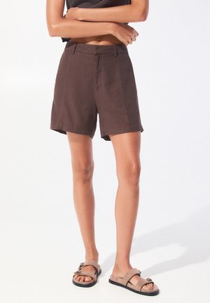 Personne portant un short taille haute marron foncé et des sandales à brides couleur sable, debout les bras croisés devant un fond blanc uni.