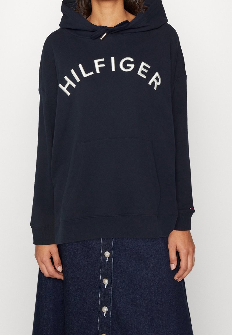 Sweat à capuche bleu marine avec une poche avant et un cordon de serrage, arborant le mot "HILFIGER" brodé en blanc. Associé à une jupe en denim avec des boutons décoratifs.