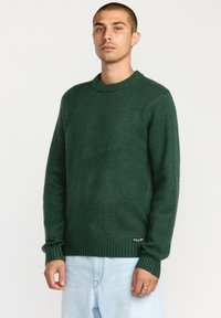 Pull en maille vert avec un col rond, des poignets et un ourlet côtelés, et une finition texturée. Petit logo en bas à gauche.