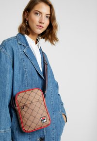 Sac bandoulière FILA marron avec un motif logo sur toute la surface, bordure rouge et bandoulière rayée réglable. Texture lisse, forme rectangulaire, poche zippée à l'avant.