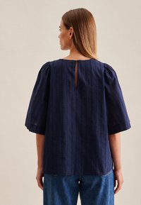 Top blu navy con maniche corte a sbuffo; presenta righe verticali e un dettaglio a chiave sul retro con chiusura a bottone. Tessuto strutturato.