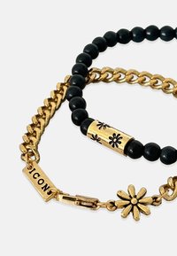 Zelta ķēde un melno krellu aproce ar dekoratīvu zelta zieda akcentu un gravētu zieda dizainu uz zelta cilindriem.